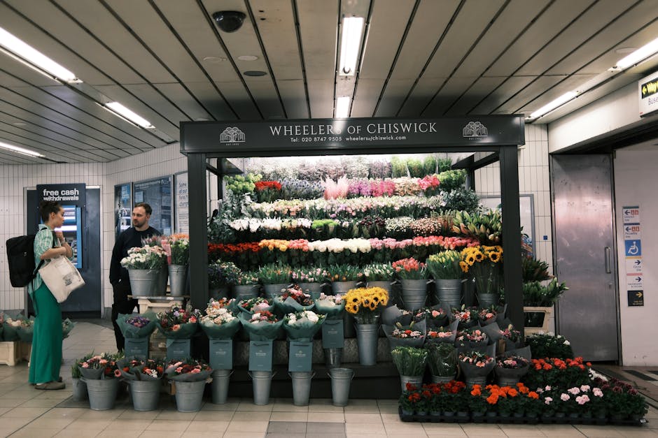 A vibrant outdoor floral display by Wheeler of Chiswick at East Finchley tube station features a variety of fresh flowers arranged in gray metal buckets and wrapping papers. The arrangement includes d